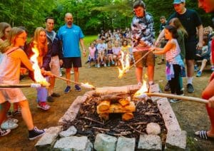Campers, staff, and director roasting marshmallows by the campfire