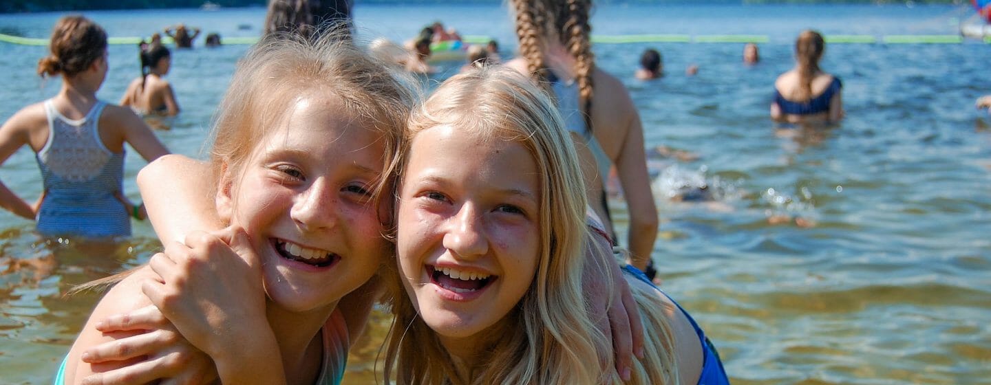 Two girls hugging each other at the waterfront with campers boating and swimming behind them